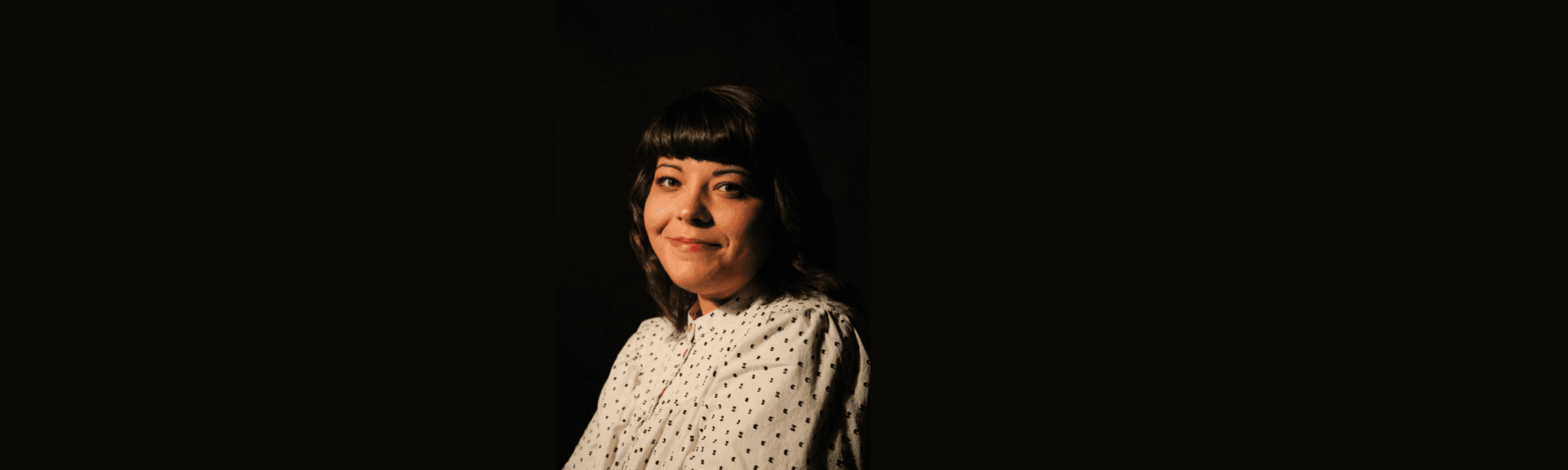 Woman with dark hair and a white patterned blouse smiling against a dark background.